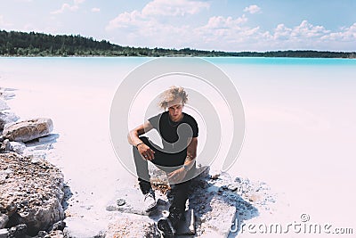 Curly blond man posing at the clear beach. Dressed all black. Awesome background, clear water and sky. Trendy, tropical and Stock Photo