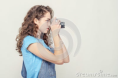 Curly brunette girl in denim overalls is holding a vintage film camera in her hands. Takes a picture. Side view Stock Photo