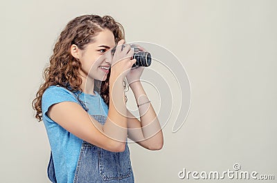 Curly brunette girl in denim overalls is holding a vintage film camera in her hands. Takes a picture. Side view Stock Photo