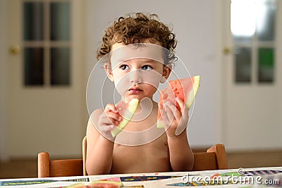 Curly-haired baby girl eats fruit sitting indoors at home in summer Stock Photo