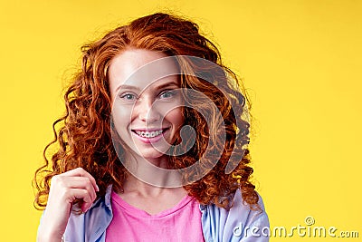 Curly redhaired ginger woman with snow white smile brackets on teeth touching a new hairstyle in studio yellow Stock Photo