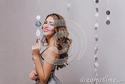 Curly stylish lady in shining dress coquettishly looks into camera on white background with silver glitter. Stock Photo