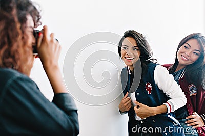 Curly tanned female photographer with red manicure taking photo of glad latin and asian girls in sport jackets. Adorable Stock Photo