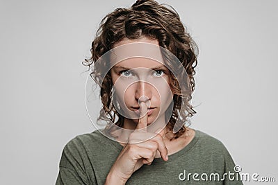 Curly caucasian young woman looks serious, holding finger at her lips asking shut up Stock Photo