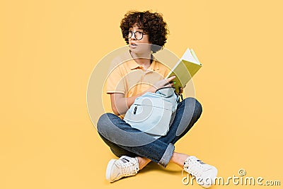 Curtly teenager with backpack holding book while sitting on yellow background. Stock Photo