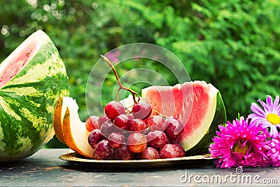 Cut slices of ripe yellow melon, watermelon, a bunch of grapes and flowers asters on a table with natural green background Stock Photo