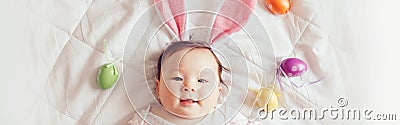 Cute adorable Asian baby wearing pink Easter bunny ears. Infant kid lying on bed with coloredl Easter eggs. Funny child Stock Photo