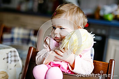 Cute adorable baby girl playing with first doll. Beautiful toddler child sitting in high chair at home. Happy healthy Stock Photo