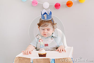 Cute adorable Caucasian baby boy in blue crown celebrating his first birthday at home. Child kid toddler sitting in a high chair Stock Photo