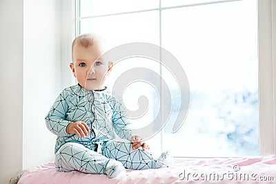 Cute adorable little baby girl sitting by window and looking to cam. Kid enjoy snowfall. Happy holidays and Christmas! Winter Home Stock Photo