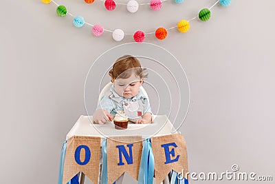 Cute adorable sad upset Caucasian baby boy celebrating his first birthday at home. Pensive child kid toddler sitting in high chair Stock Photo