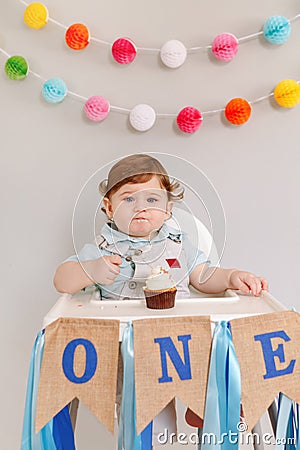Cute adorable sad upset Caucasian baby boy celebrating his first birthday at home. Pensive child kid toddler sitting in high chair Stock Photo