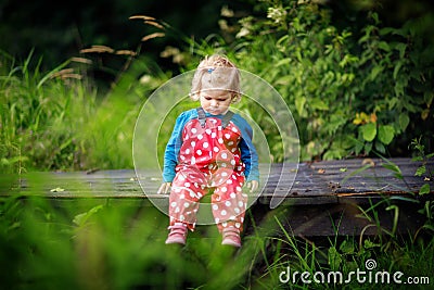 Cute adorable toddler girl sitting on wooden bridge and throwing small stones into a creek. Funny baby having fun with Stock Photo