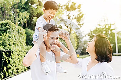 Cute Asian father piggybacking his son along with his wife in the park. Excited family spending time together with happiness. Stock Photo