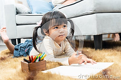 A cute Asian girl is lying on the carpet on the floor of the house with a crayon in her hand happily drawing on a notebook in the Stock Photo