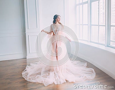 Cute attractive princess with blond hair in pink light dress posing in sunlight of large window, sleeping beauty woke up Stock Photo