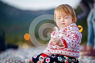 A cute baby in a beautiful sweater on the shore of the winter sea plays with stones. The kid sits in profile on a stone beach. Stock Photo
