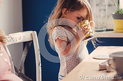 Cute baby girl drinking tea for breakfast in sunny kitchen Stock Photo