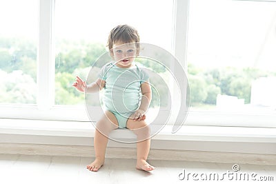 Cute baby at home in white room is sitting near window. The beautiful baby could be a boy or girl and is wearing body suit. Stock Photo