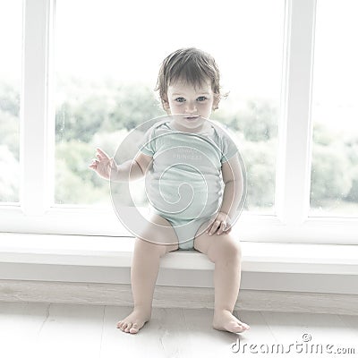 Cute baby at home in white room is sitting near window. The beautiful baby could be a boy or girl and is wearing body suit. Stock Photo