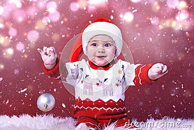 Cute baby in Santa hat with Christmas ball on fluffy carpet against red background. Magical festive atmosphere Stock Photo