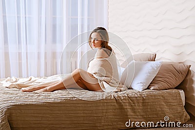 Cute barefoot woman sits on bed wrapped in blanket Stock Photo