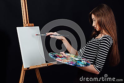Cute beautiful girl artist painting a picture on canvas an easel. Space for text. Studio black background. Stock Photo
