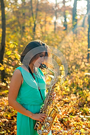 Cute black-haired girl in a blue dress plays the yellow saxophone in the autumn forest Stock Photo