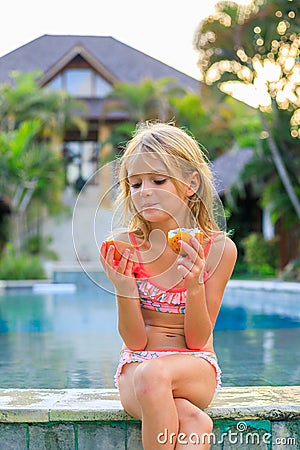 Cute blond little girl in pink swimsuit sitting at the swimming pool, holding passion fruit. Enjoy eating tropical fruit. Summer Stock Photo