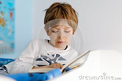 Cute blond little kid boy in pajamas reading book in his bedroom. Excited child reading loud, sitting in his bed Stock Photo