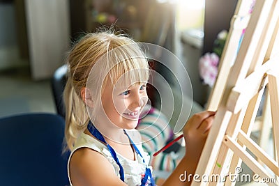 Cute blond smiling caucasian kid painting on wooden easel in class workshop lesson at art studio. Little girl holding brush in Stock Photo