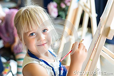 Cute blond smiling girl painting on easel in workshop lesson at art studio. Kid holding brush in hand and having fun drawing with Stock Photo