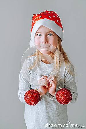 Cute blonde toddler girl wearing a santa hat holding two red Christmas balls of Christmas tree decoration against Stock Photo