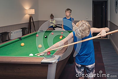 Cute boy in blue t shirt plays billiard or pool in club. Young Kid learns to play snooker. Boy with billiard cue strikes the ball Stock Photo