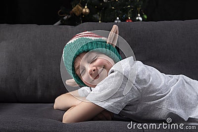 Cute boy in christmas elf hat is resting on the sofa with closed eyes on the background of Christmas tree. Anticipation of holiday Stock Photo