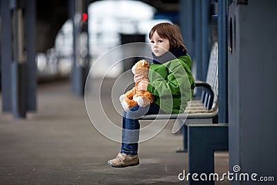 Cute boy, sitting on a bench with teddy bear, looking at a train Stock Photo