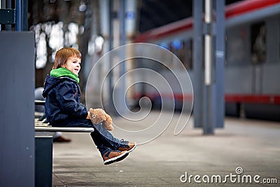 Cute boy, sitting on a bench with teddy bear, looking at a train Stock Photo