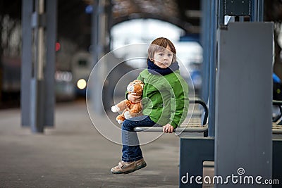 Cute boy, sitting on a bench with teddy bear, looking at a train Stock Photo