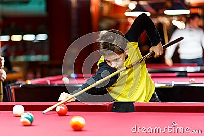 Cute boy in yellow t shirt plays billiard or pool in club. Young Kid learns to play snooker. Boy with billiard cue Stock Photo