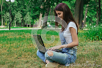 Cute brunette sits in a park on the lawn and prints on a laptop Stock Photo