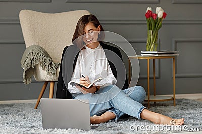 A cute business lady is sitting in front of a laptop on a gray carpet on the floor, wearing a white shirt with a black jacket on Stock Photo