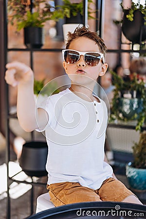 Cute cauasian boy in white tee shirt and sunglasses sitting at the table in loft style cafe and pointing somewhere. Stock Photo