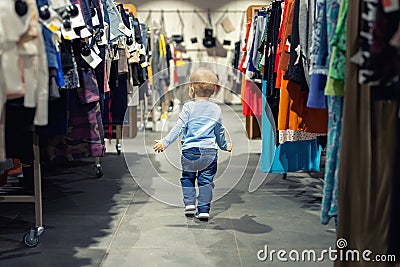 Cute caucasian blond toddler boy walking alone at clothes retail store between rack with hangers. Baby discovers adult shopping Stock Photo