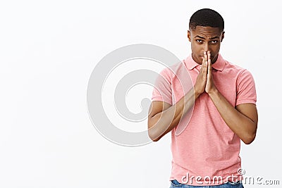 Cute and charismatic african american guy in pink polo shirt bowing holding hands in pray against face peeking from Stock Photo