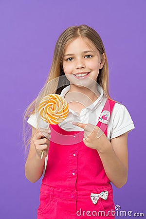 Cute cheerful girl holding giant candy. Kid with long blond hair isolated on violet background. Child pointing at Stock Photo