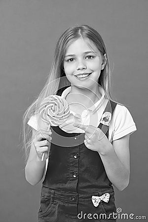 Cute cheerful girl holding giant candy. Kid with long blond hair isolated on violet background. Child pointing at Stock Photo