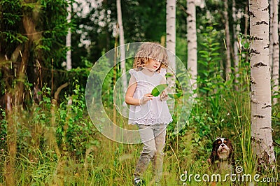 Cute child girl playing with leaves in summer forest with her dog. Nature exploration with kids. Stock Photo