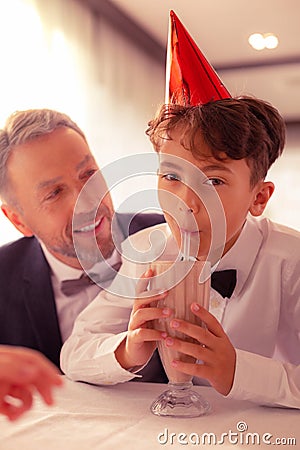 Cute dark-eyed grandson drinking milk cocktail near his granddad Stock Photo