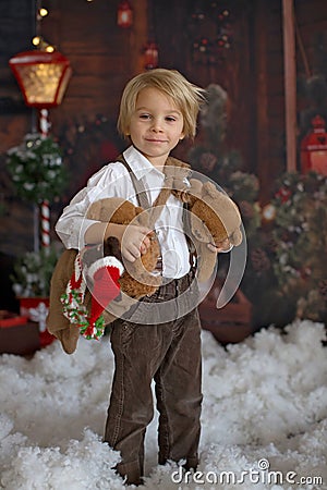 Cute fashion toddler boy, playing in the snow with teddy bear in front of a wooden cabin log Stock Photo