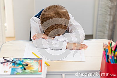 A cute first grader boy in a school uniform at home during a pandemic fell asleep doing homework at a desk with books and pencils. Stock Photo